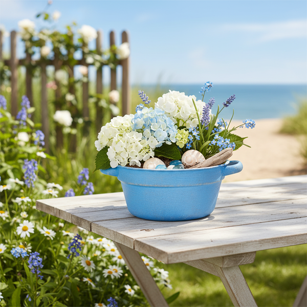 Blumenarrangement mit Hortensien und Lavendel in blauem Pflanzgefäß mit Muscheln auf einem Holztisch im Garten mit Meerblick.