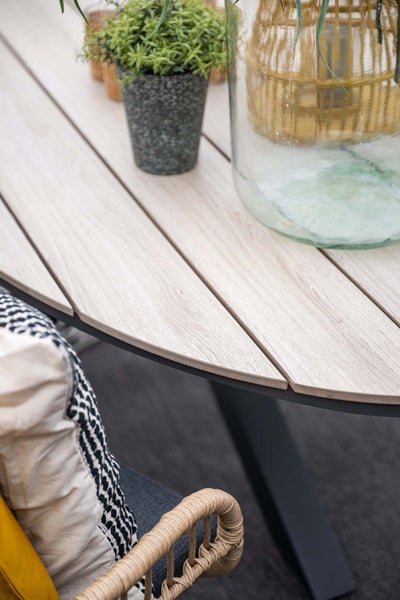 Detailansicht eines runden Gartentisches mit hellen Lamellen in Holzoptik und dunklem Metallgestell, dekoriert mit Pflanze und Glasvase.