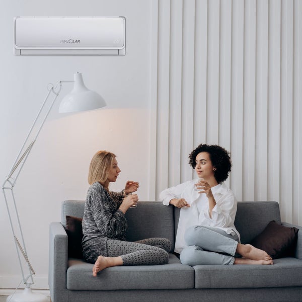FM-SOLAR Klimaanlage an einer Wand in einem hellen Wohnzimmer mit zwei Frauen auf einem grauen Sofa und einer Stehlampe.