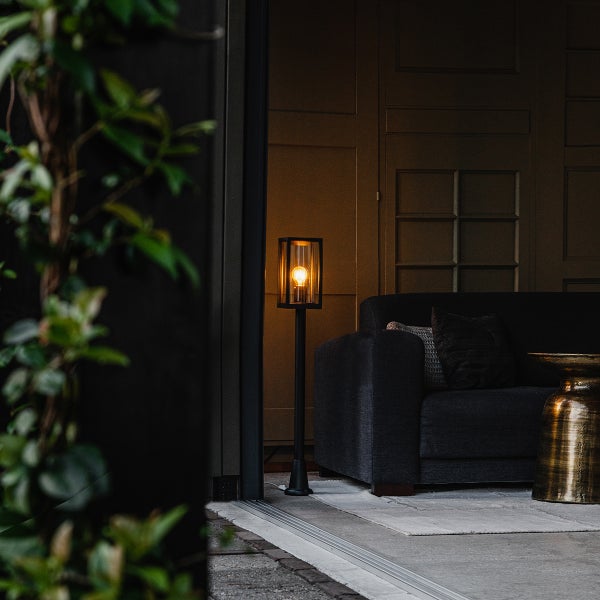 Moderne schwarze Außenstehleuchte mit Glaslaterne auf einer Terrasse mit dunklem Sofa und goldenem Beistelltisch.