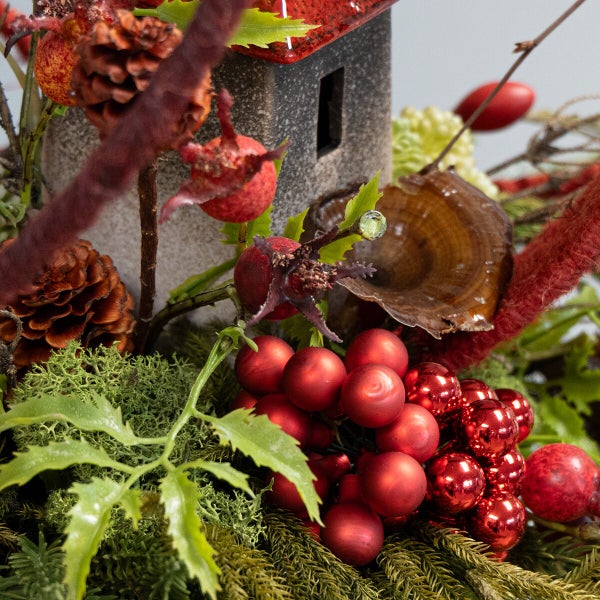 Weihnachtliche Tischdekoration mit Keramikhaus, Tannenzapfen, Moos, künstlichen Beeren und glänzenden Christbaumkugeln.
