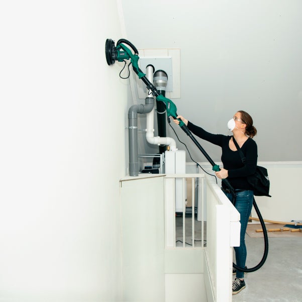 Person schleift eine Wand mit einem Langhalsschleifer in einem Innenraum mit einer Atemschutzmaske