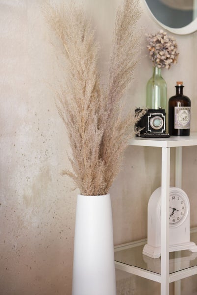 Eine weiße Vase mit getrocknetem Pampasgras neben einem Regal mit einer Vintage-Kamera, Glasflaschen und einer weißen Tischuhr.