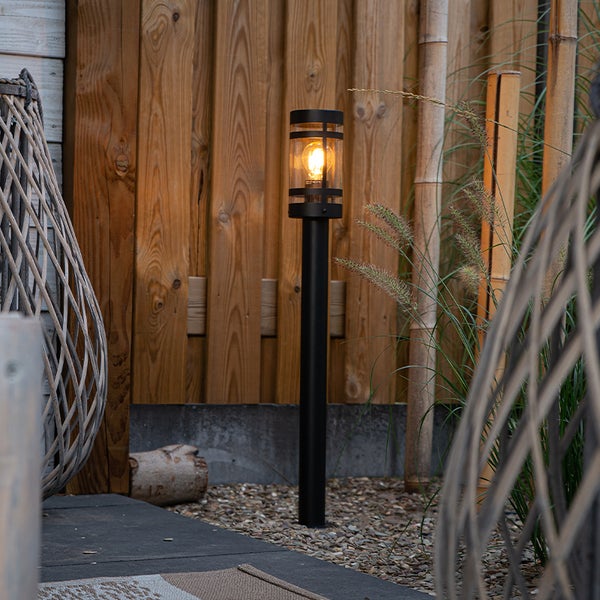 Außen-Stehleuchte in Schwarz mit leuchtendem Leuchtmittel vor einem Holzzaun im Garten.