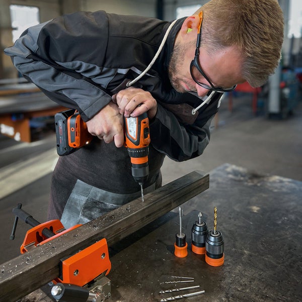 Ein Mann bohrt mit einer Akku-Bohrmaschine ein Metallstück, das mit einer Schraubzwinge fixiert ist. Verschiedene Bohrer liegen bereit.