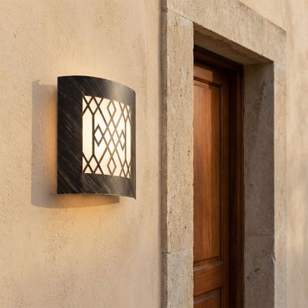 Außenwandleuchte aus dunklem Metall mit geometrischem Gittermuster und warmem Licht, montiert an einer hellen Hauswand neben einer Holztür.