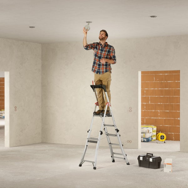 Mann auf einer Trittleiter, der eine Glühbirne in einem Raum installiert. Werkzeugkasten und Baumaterialien sind im Raum.