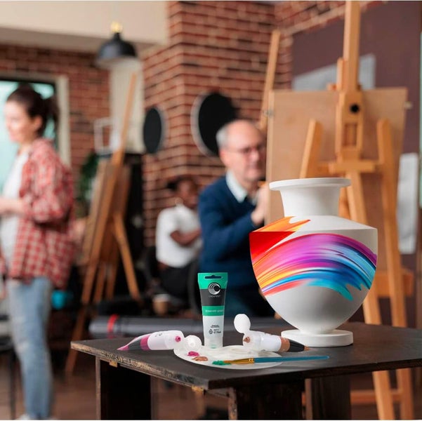 Bemalte Keramikvase mit bunten Pinselstrichen auf einem Holztisch neben Acrylfarbtuben und einer Palette in einem Kunstatelier.