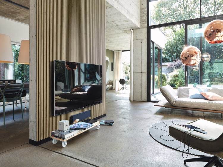 Modernes Wohnzimmer mit Lamellenwand aus Holz und wandmontiertem Fernseher sowie großen Fensterfronten und Sichtbeton.
