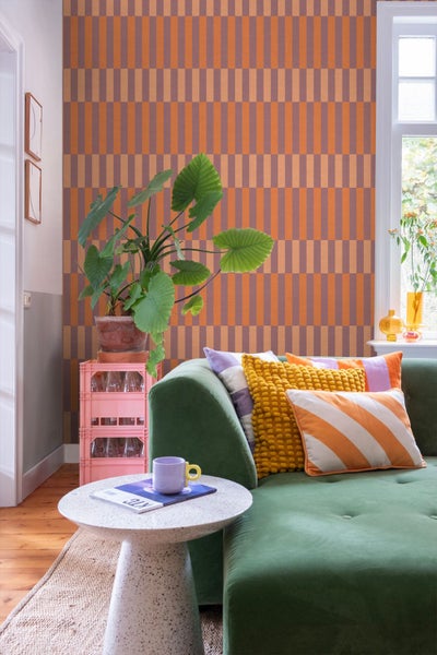 Wohnzimmer mit geometrisch gemusterter Tapete, einem grünen Sofa mit bunten Kissen und einer großen Zimmerpflanze.
