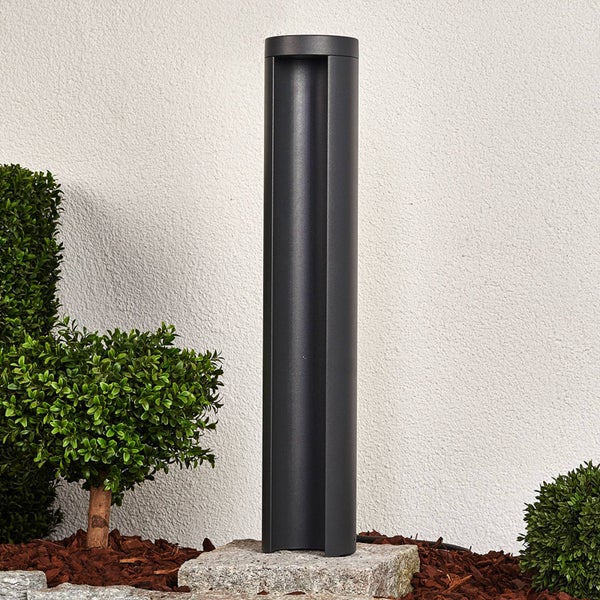 Moderne zylindrische Wegeleuchte in Anthrazit auf einem Steinsockel im Garten mit kleinen grünen Sträuchern.