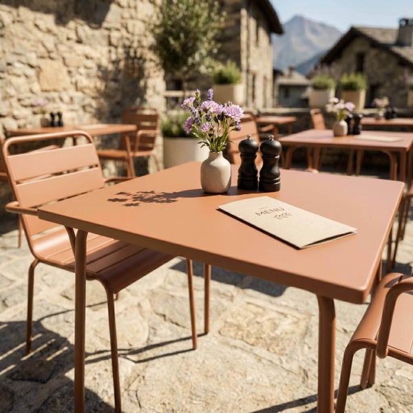 Terracottafarbener Metalltisch und Stühle auf einer rustikalen Steinterrasse mit einer kleinen Blumenvase, einer Menükarte und Gewürzstreuern.