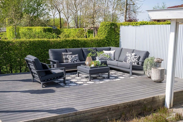 Moderne Garten-Lounge-Gruppe mit dunkelgrauem Ecksofa, Sessel und Couchtisch auf einer Holzterrasse mit Outdoorteppich vor einer grünen Hecke.