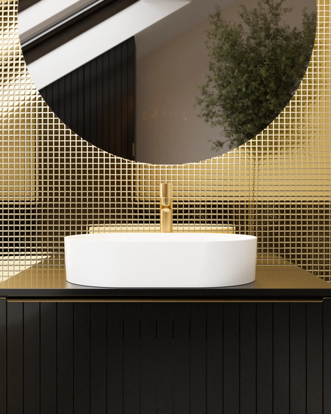 Modernes Badezimmer mit weißem Aufsatzwaschbecken auf dunklem Unterschrank vor einer Wand aus goldenen Mosaikfliesen und kreisförmigem Spiegel.