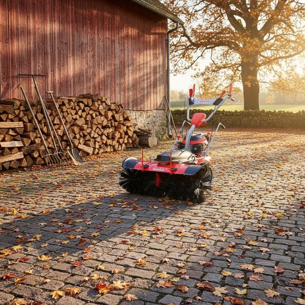 Kehrmaschine im Außeneinsatz auf Pflastersteinen mit Laub, neben einem Holzstapel und Gartengeräten