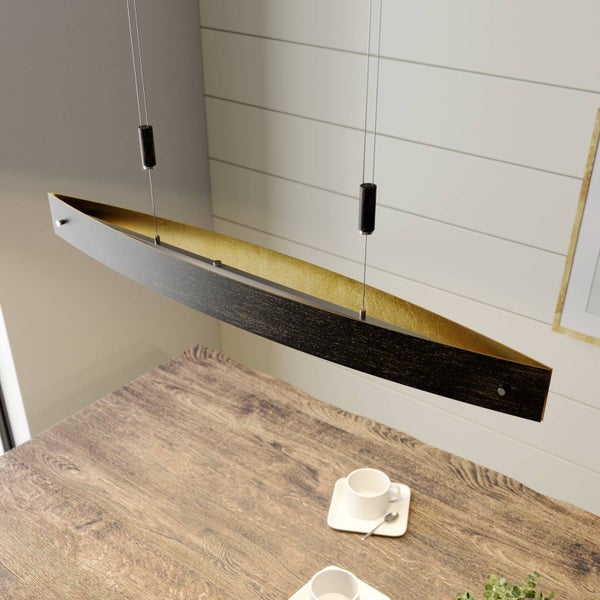 Moderne Pendelleuchte in länglicher, gebogener Form über einem Holztisch. Außen schwarz strukturiert, innen goldfarben.
