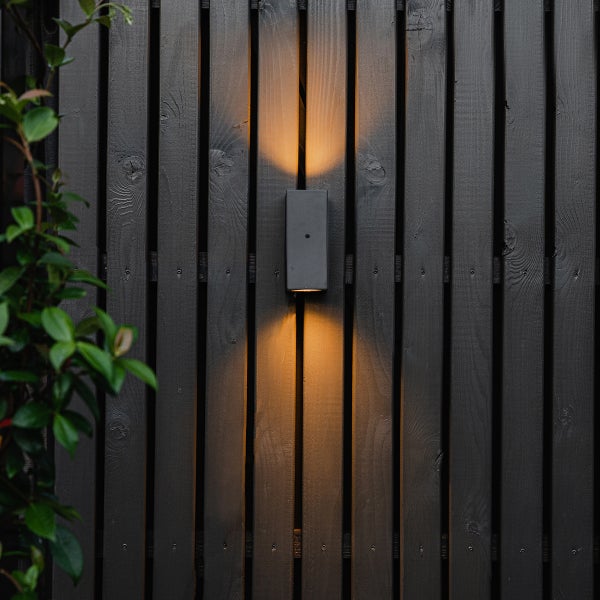 Moderne rechteckige Außenwandleuchte an einem dunklen Holzzaun mit warmem Lichtschein nach oben und unten.