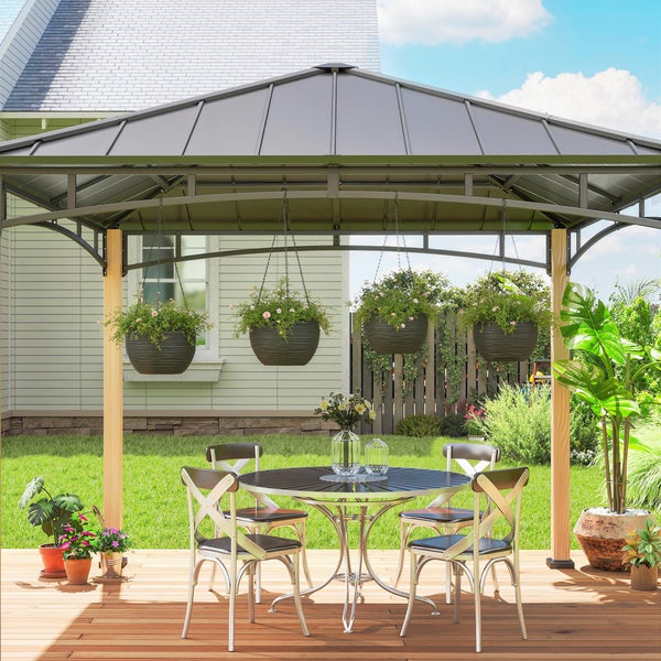Moderner Metallpavillon mit festem Walmdach und Holzpfosten über einem runden Tisch mit Stühlen auf einer Holzterrasse in einem hellen Garten.