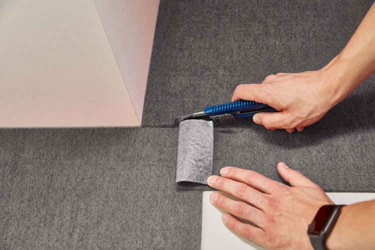 Eine Person schneidet graues Vlies mit einem blauen Cuttermesser auf einer ebenen Fläche zurecht.
