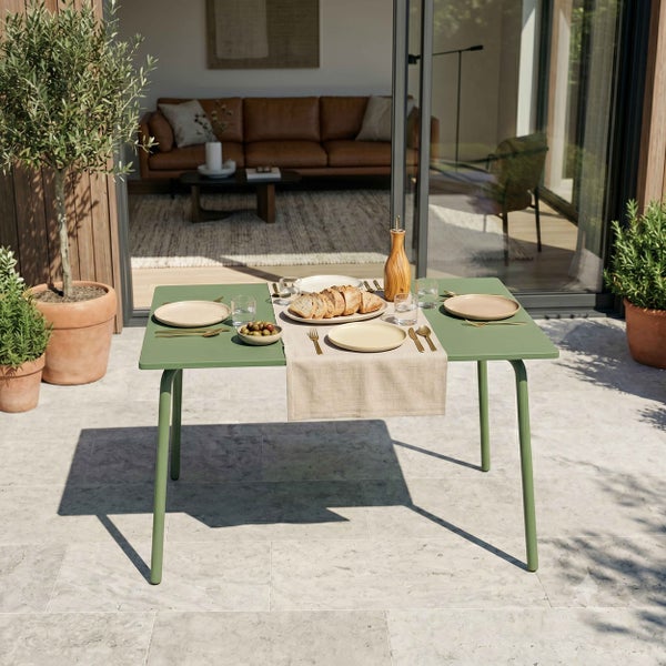Gedeckter grüner Gartentisch auf einer hellen Terrasse aus Steinplatten vor einer Glasfront mit Blick in ein Wohnzimmer.