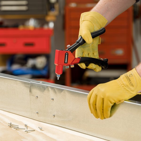 Eine Person mit gelben Arbeitshandschuhen bedient eine Blindnietzange von Arrow an einem Metallprofil in einer Werkstatt.