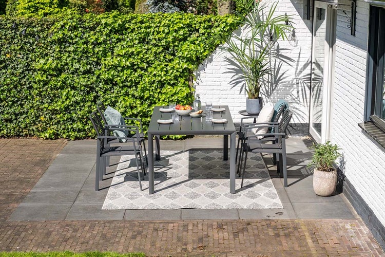 Modernes Gartenmöbel-Set mit dunklem Metalltisch und vier Stühlen auf einer gepflasterten Terrasse mit Außenteppich vor einer grünen Hecke.