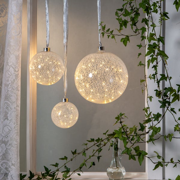 Dekorative, leuchtende Weihnachtskugeln hängen an Bändern vor einem Fenster mit Efeu.