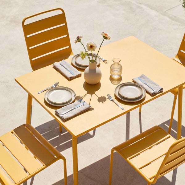 Modernes Gartenmöbel-Set aus gelbem Metall mit einem rechteckigen Tisch und vier Stühlen in Lamellenoptik, gedeckt auf einer sonnigen Terrasse.
