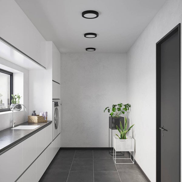 Heller, moderner Hauswirtschaftsraum mit weißen Schränken, dunkler Arbeitsplatte, drei schwarzen runden Deckenleuchten und dunklen Bodenfliesen.