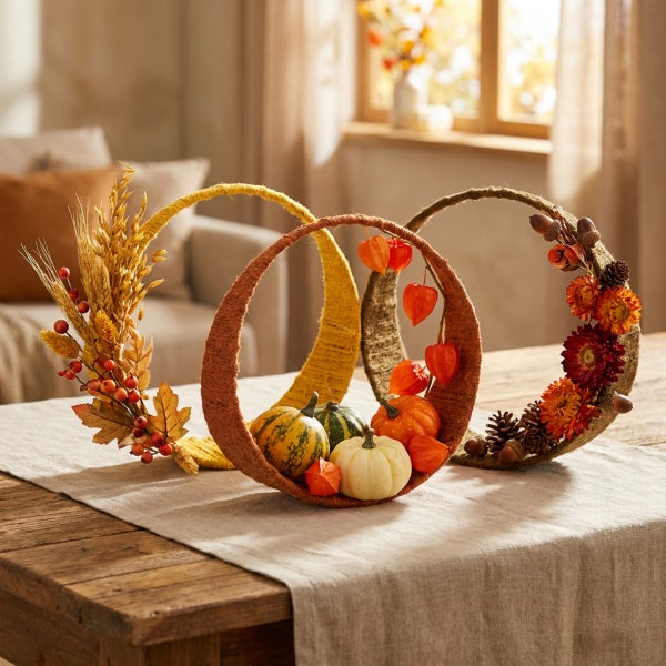 Herbstliche Tischdekoration mit drei verbundenen, umwickelten Ringen, verziert mit Kürbissen, getrockneten Blumen, Beeren und Zapfen auf einem Tisch.