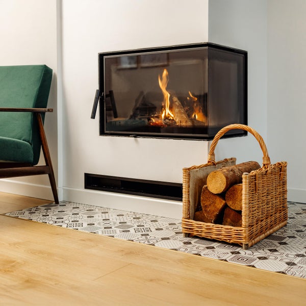 Moderner Eckkamin mit brennendem Feuer und Holzscheiten, daneben ein Weidenkorb mit Brennholz auf einer gemusterten Bodenschutzplatte.