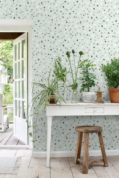 Dekorative Tapete mit Terrazzo-Muster hinter einem weißen Tisch mit Pflanzen und einem Holzhocker