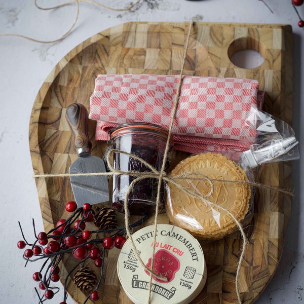 Geschenkset mit Käse, Marmelade, Waffeln, Käsemesser und Geschirrtuch auf Holzbrett