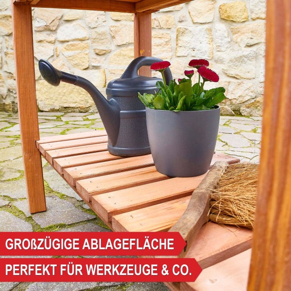 Hochbeet aus Holz mit Kräutern, untere Ablage mit Gartenwerkzeug, auf einer Terrasse vor einer Ziegelwand mit Rosen, hell und organisiert.