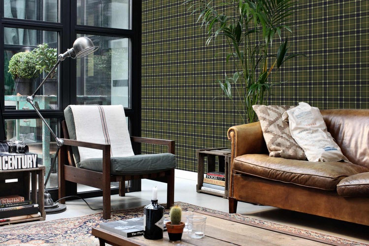 Gemütliches Wohnzimmer mit grün karierter Vliestapete im Tartan-Muster, Ledersofa, Holzsessel und Stehlampe im Industrial-Design.