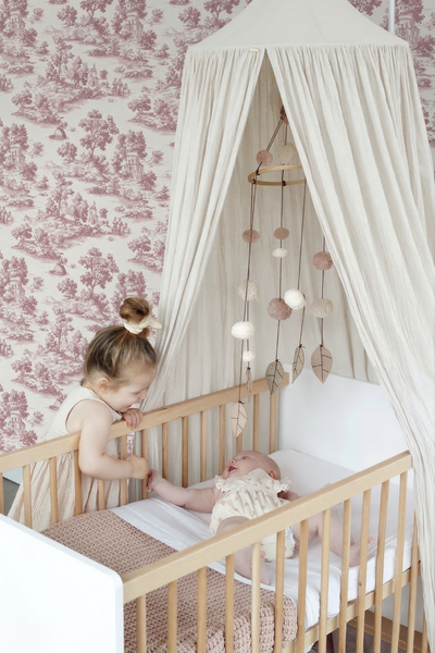 Helles Kinderzimmer mit Gitterbett aus Naturholz, weißem Betthimmel, Mobile und floraler Tapete.