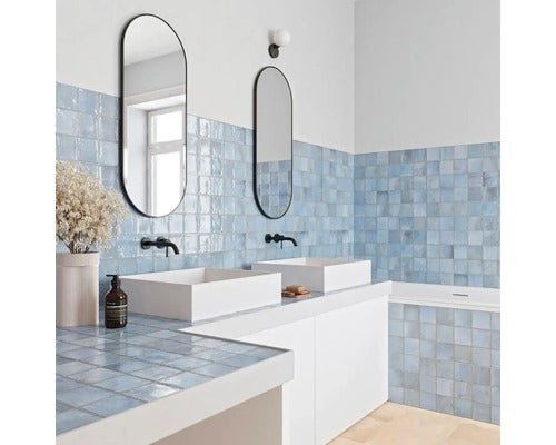 Modernes Bad mit hellblauen glänzenden quadratischen Wandfliesen, zwei weißen Aufsatzwaschbecken, schwarzen Armaturen und ovalen Spiegeln.