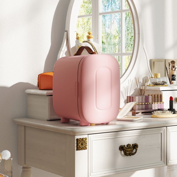 Kleiner rosafarbener Kosmetikkühlschrank auf einem weißen Schminktisch mit Spiegel und Make-up-Artikeln in einem hellen, aufgeräumten Zimmer.