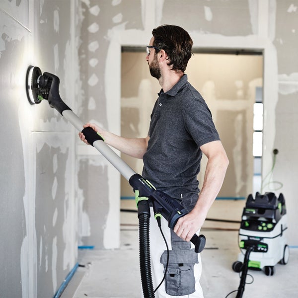 Ein Mann verwendet einen Langhalsschleifer von Festool zum Schleifen einer Wand. Ein Staubsauger ist im Hintergrund zu sehen.