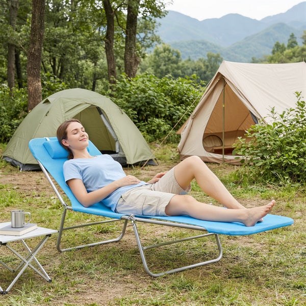 Eine Frau entspannt auf einer blauen klappbaren Sonnenliege beim Camping mit Zelten und Bergen im Hintergrund.