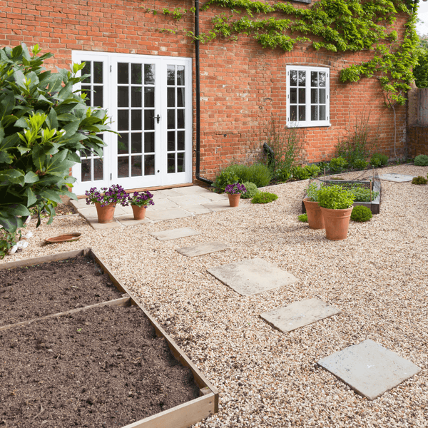Gartenanlage vor einem Backsteinhaus mit Kiesweg, Trittplatten, Hochbeeten und Topfpflanzen.