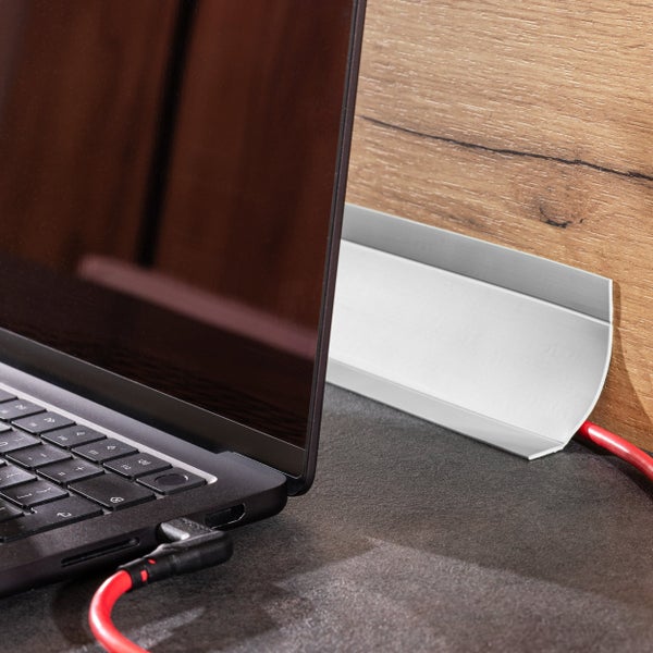 Eine silberne Kabelkanal Leiste an einer Wand aus Holzoptik neben einem Laptop auf einem Tisch mit einem roten Kabel.