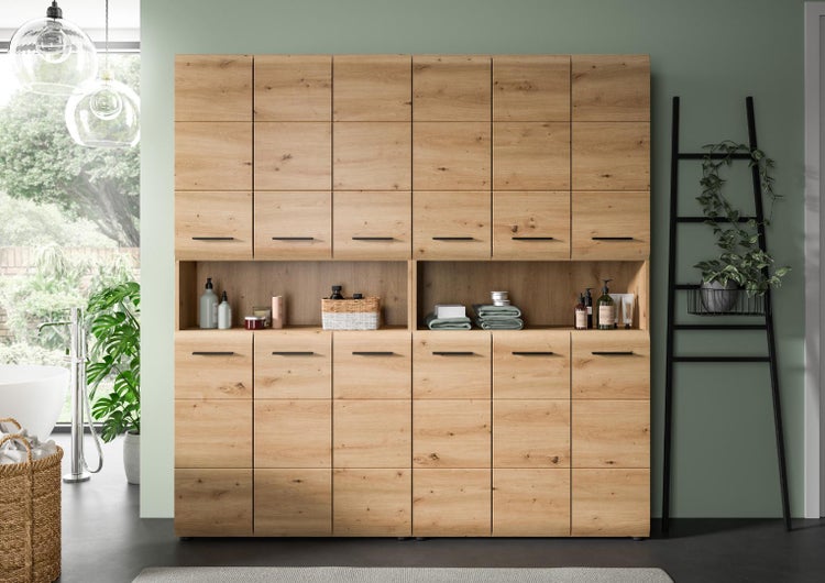 Große Badezimmerschrank-Wand in Holzoptik mit zahlreichen Fächern, schwarzen Griffen und offener Ablage in einem modernen, hellen Badezimmer.