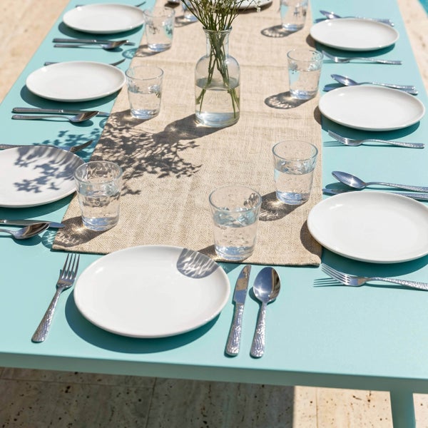 Gedeckter Gartentisch mit weißen Tellern, silbernem Besteck in Hammerschlag-Optik, Trinkgläsern und Tischläufer aus Jute auf einem türkisfarbenen Tisch.