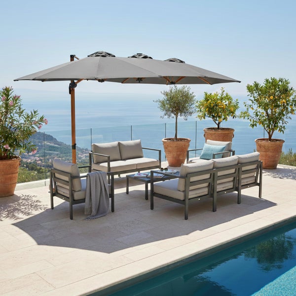 Moderne Gartenlounge aus Metall mit grauen Polstern, großem Ampelschirm mit Holzmast auf einer Steinterrasse am Pool mit Meerblick.