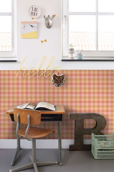 Hell gestaltetes Kinderzimmer mit karierter Tapete in Rosa- und Orangetönen, einem hölzernen Schreibtisch, Stuhl und verspielter Wanddekoration.