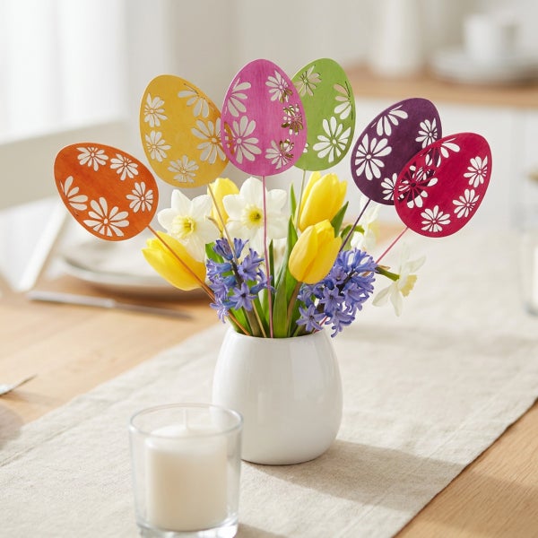 Osterstrauß in einer weißen Vase mit Tulpen, Narzissen, Hyazinthen und hölzernen Ostersteckern mit Blumenmuster auf einem Tisch.