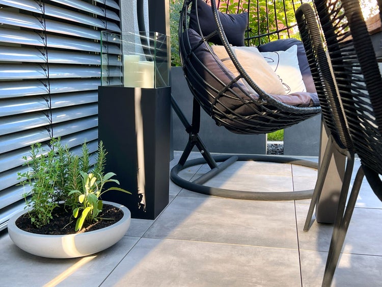 Moderne Terrasse mit Hängesessel, großer schwarzer Laterne und runder Pflanzschale mit Kräutern auf grauen Fliesen.