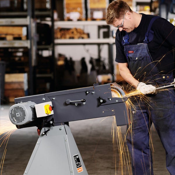 Handwerker schleift Metall an einer stationären Bandschleifmaschine mit Funkenflug und Schutzkleidung in einer Werkstatt.