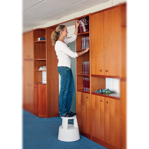 Frau steht auf Tritthocker und räumt Buch in Schrank ein